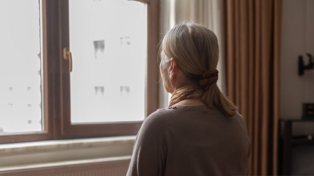 Older woman looks out the window in her apartment.