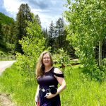 Angela Branson holds a camera and is looking at us. In the background, it is springtime in Vail, Colorado with bright green foliage everywhere and a road winding into the trees.