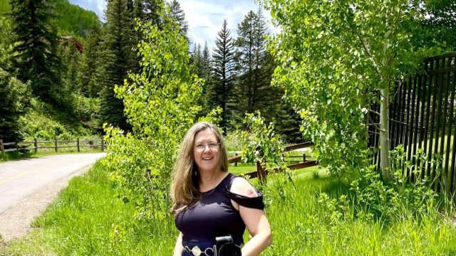 Angela Branson holds a camera and is looking at us. In the background, it is springtime in Vail, Colorado with bright green foliage everywhere and a road winding into the trees.