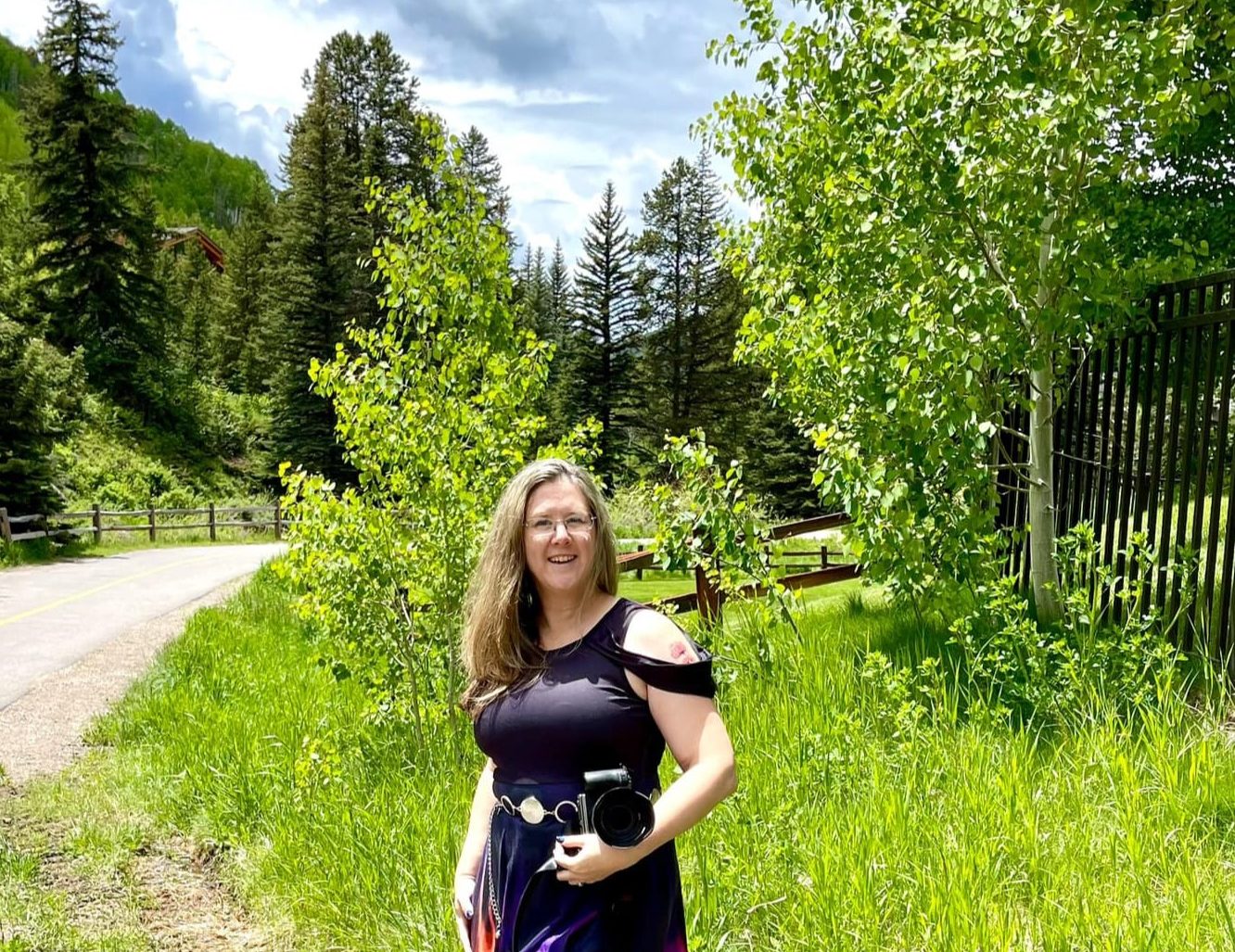 Angela Branson holds a camera and is looking at us. In the background, it is springtime in Vail, Colorado with bright green foliage everywhere and a road winding into the trees.