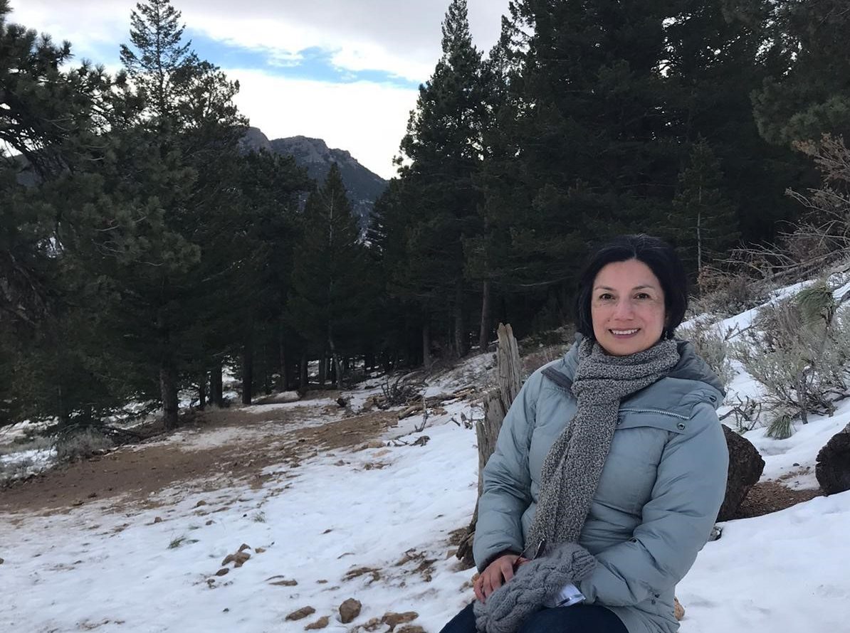 María Fernanda Enríquez Szentkirályi sits on a rock with a view of the mountains and pine trees in the background. Snow is on the ground, and she wears a light blue puffer coat.
