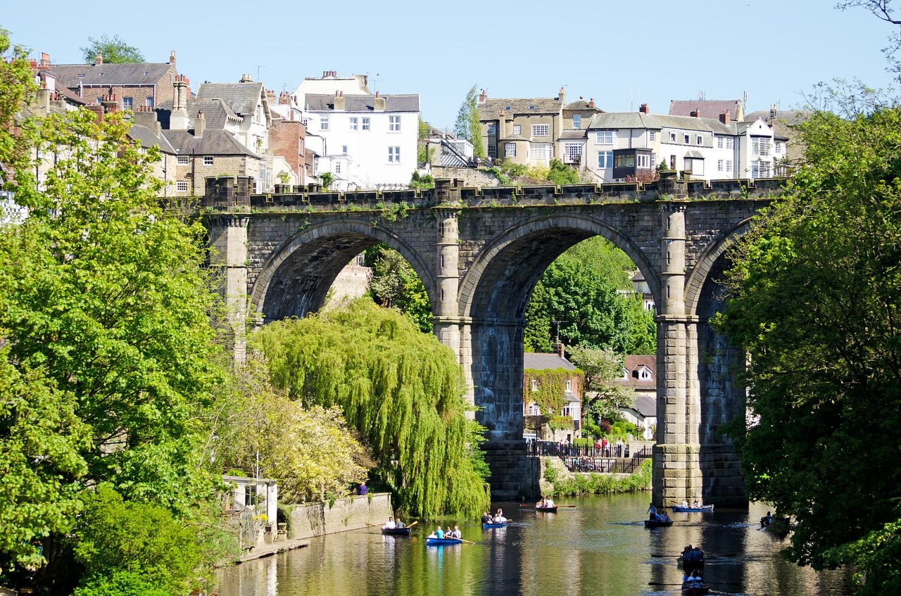 A river runs through a tall bridge with arches, reminiscent of Roman aqueducts. Across the river is a small English village. People are in the river on small canoes.