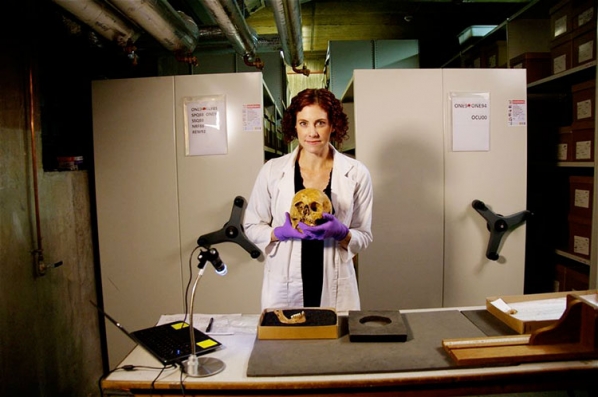 Sharon Dewitte wears a white lab coat and holds a skull with both hands. She stands near a desk.