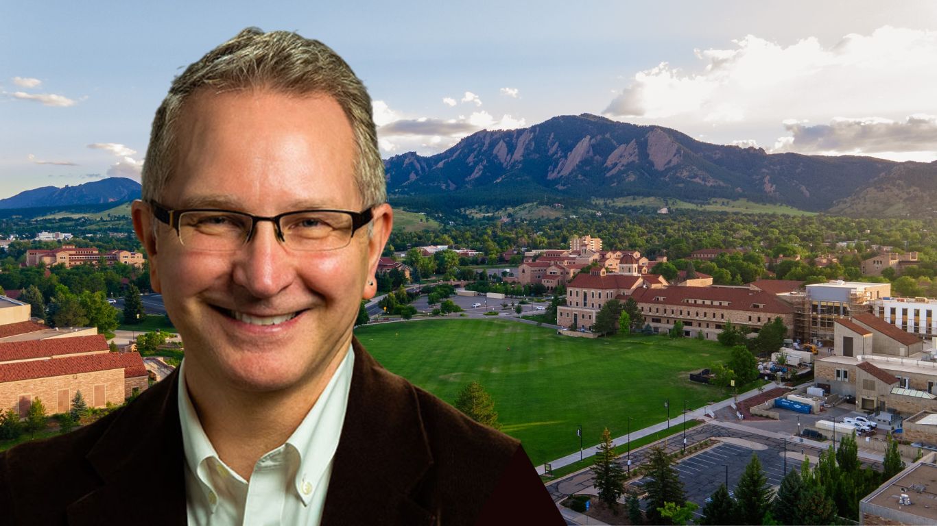 Karl Hill wears a brown jacket and black glasses. In the background are the Flatirons and CU Boulder campus.