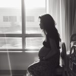 A pregnant woman sits beside her hospital bed. In the background, a large window with sunlight.