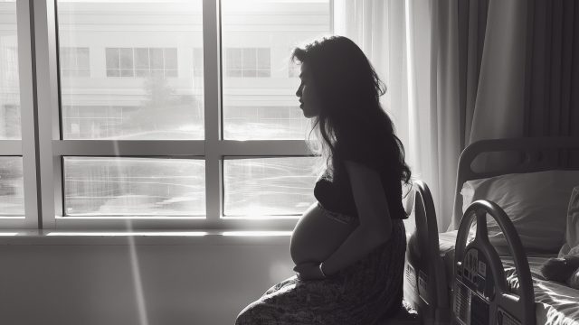 A pregnant woman sits beside her hospital bed. In the background, a large window with sunlight.