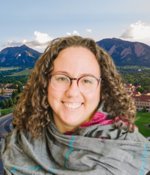 Leigh Senderowicz smiles, wearing round glasses. Behind, a view of the CU Boulder campus against the flatirons.