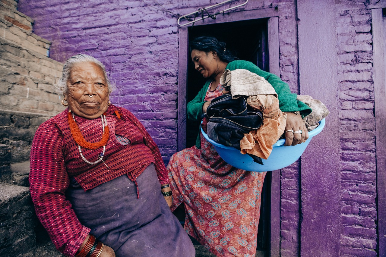 An older Nepalese woman wears a red shirt. Behind her, her daughter holds a basket of laundry. The building behind them is purple.