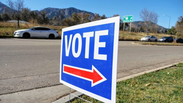 A voting sign is posted outside a voting center near the Baseline Road exit off Highway 157. Mountains can be seen in the distance as well as a white car parked on the road.