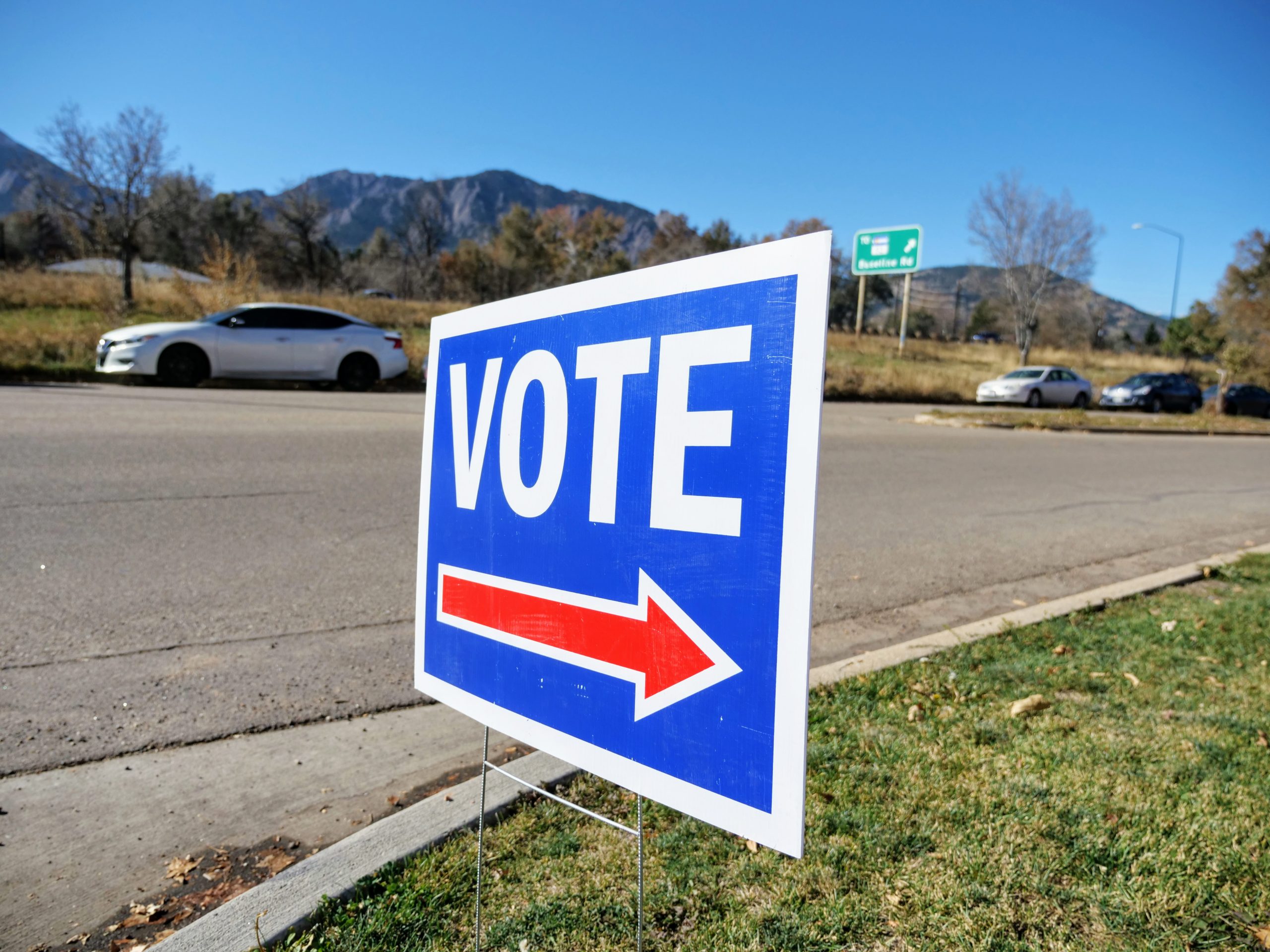 A voting sign is posted outside a voting center near the Baseline Road exit off Highway 157. Mountains can be seen in the distance as well as a white car parked on the road.