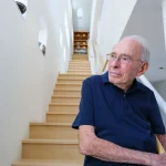 Richard Jessor wears a navy polo shirt and stands at the bottom of a stairwell.