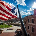 The United States Flag flaps in the breeze. Behind, a cloudy blue sky and view of the CU Boulder campus.