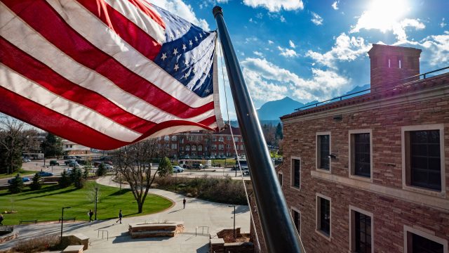 The United States Flag flaps in the breeze. Behind, a cloudy blue sky and view of the CU Boulder campus.