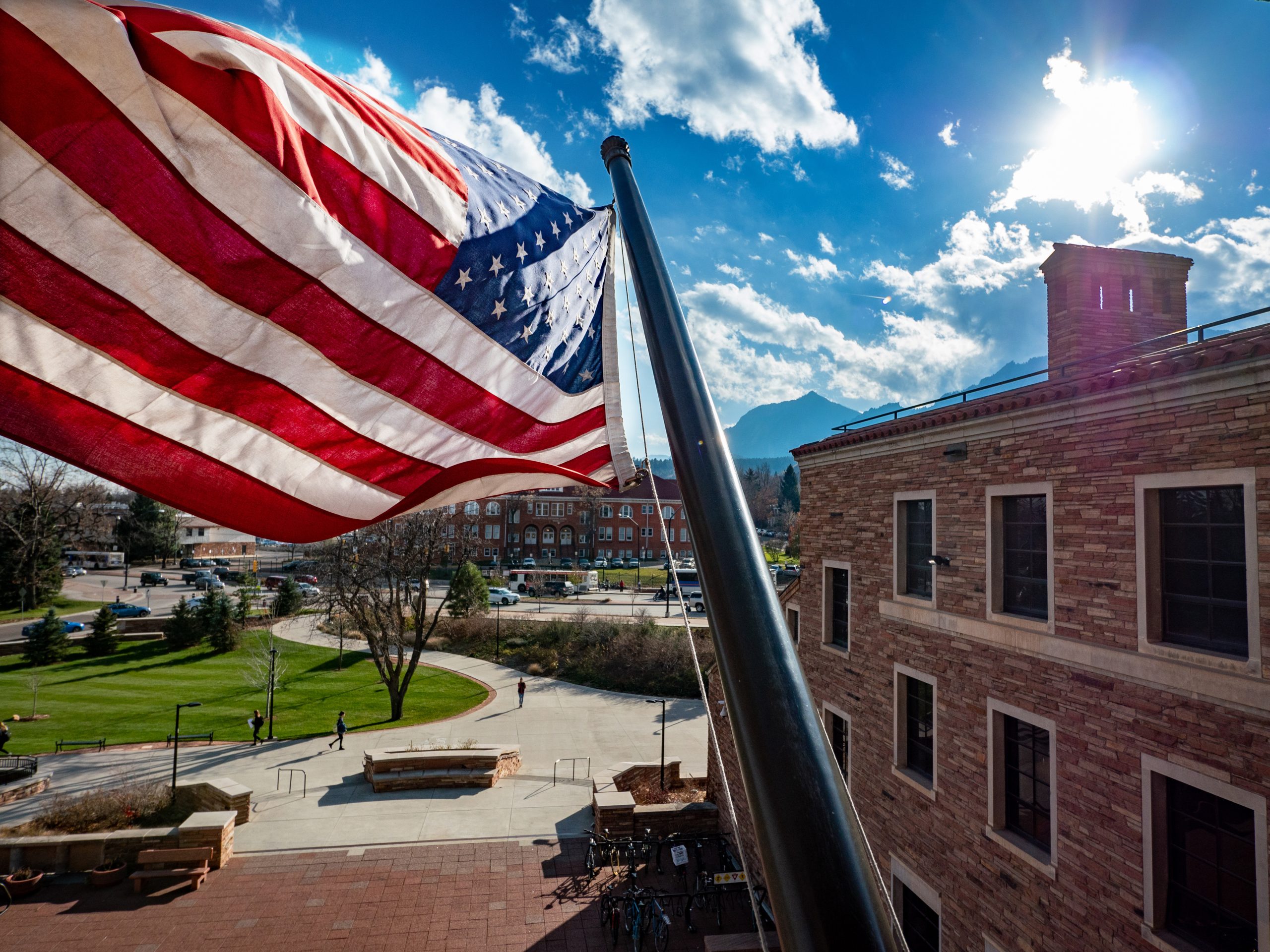 The United States Flag flaps in the breeze. Behind, a cloudy blue sky and view of the CU Boulder campus.