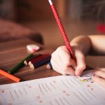 A young girl writes on a piece of paper with a red colored pencil.