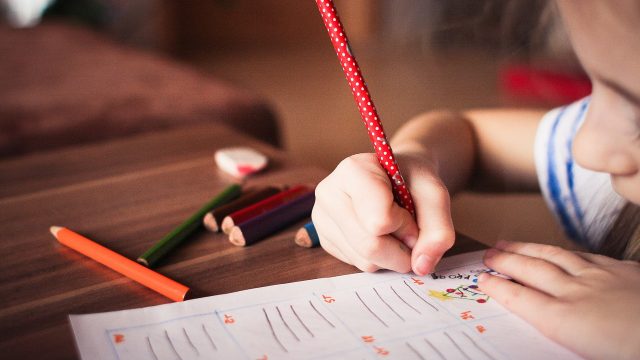 A young girl writes on a piece of paper with a red colored pencil.