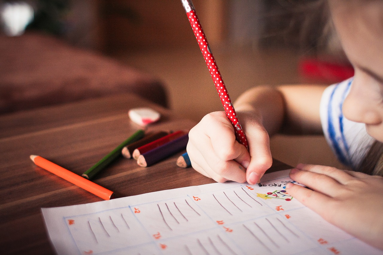 A young girl writes on a piece of paper with a red colored pencil.