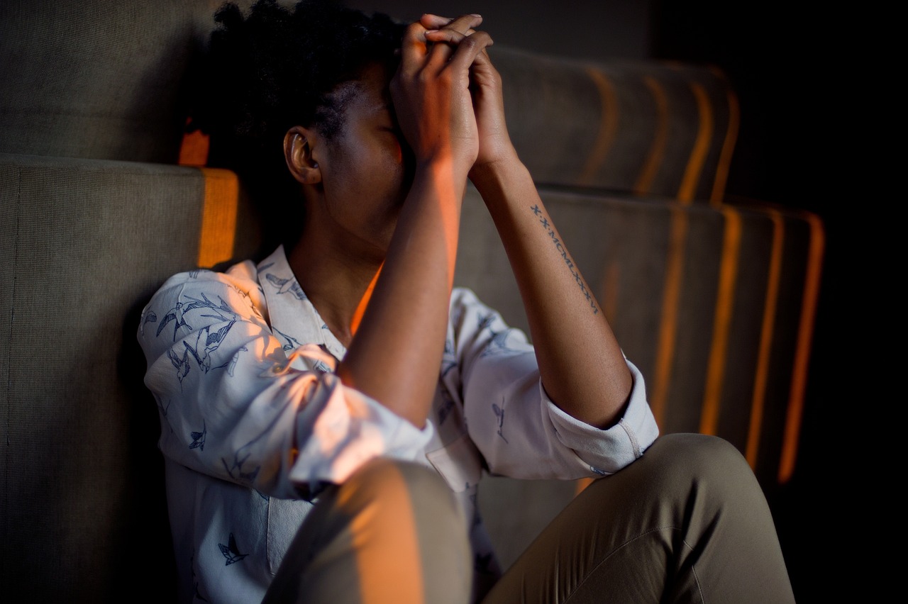 A woman sits on the stairs with her elbows on her knees, palms in a prayer position in front of her face. She has a tattoo along her left forearm.