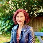 Sharon DeWitte wears a leather jacket and sits in front of greenery.