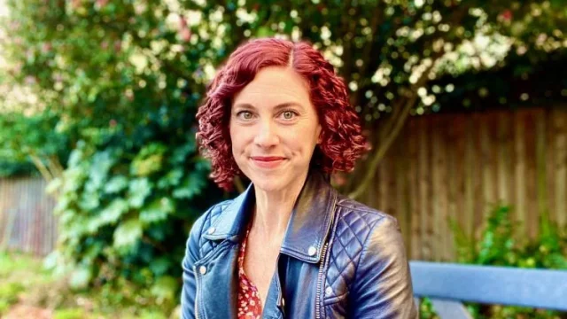 Sharon DeWitte wears a leather jacket and sits in front of greenery.