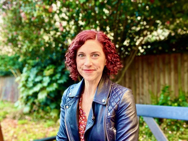 Sharon DeWitte wears a leather jacket and sits in front of greenery.