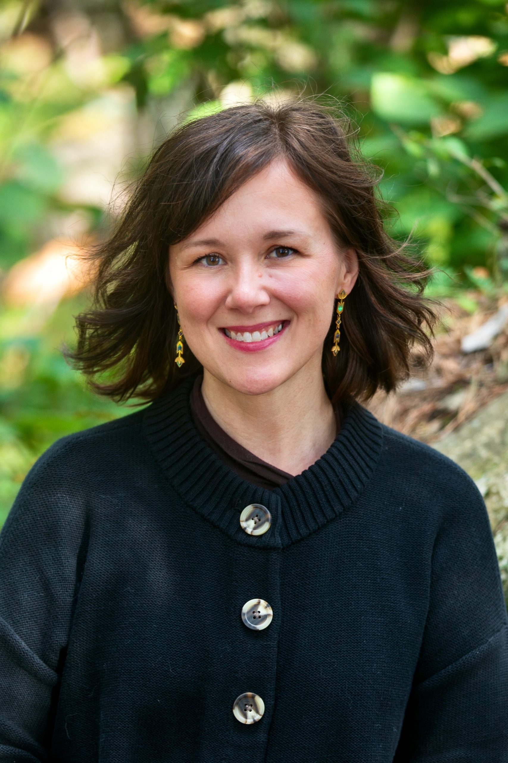 Melissa Walls wears a black jacket and sits behind some green foliage.