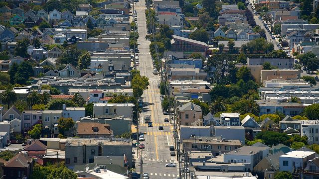 A city street evenly divides a suburban neighborhood. Trees and greenery are scattered between the homes/buildings.