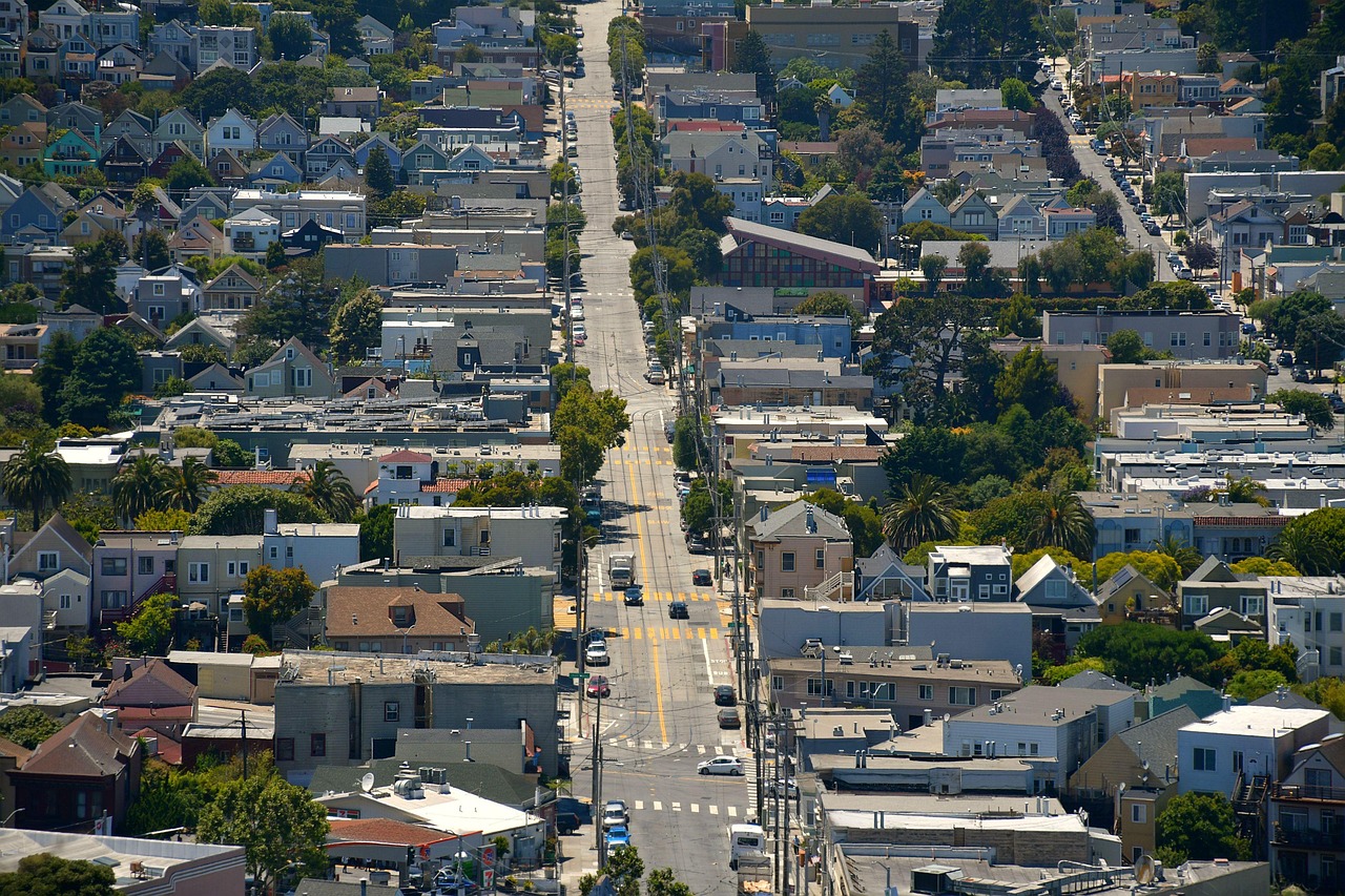 A city street evenly divides a suburban neighborhood. Trees and greenery are scattered between the homes/buildings.